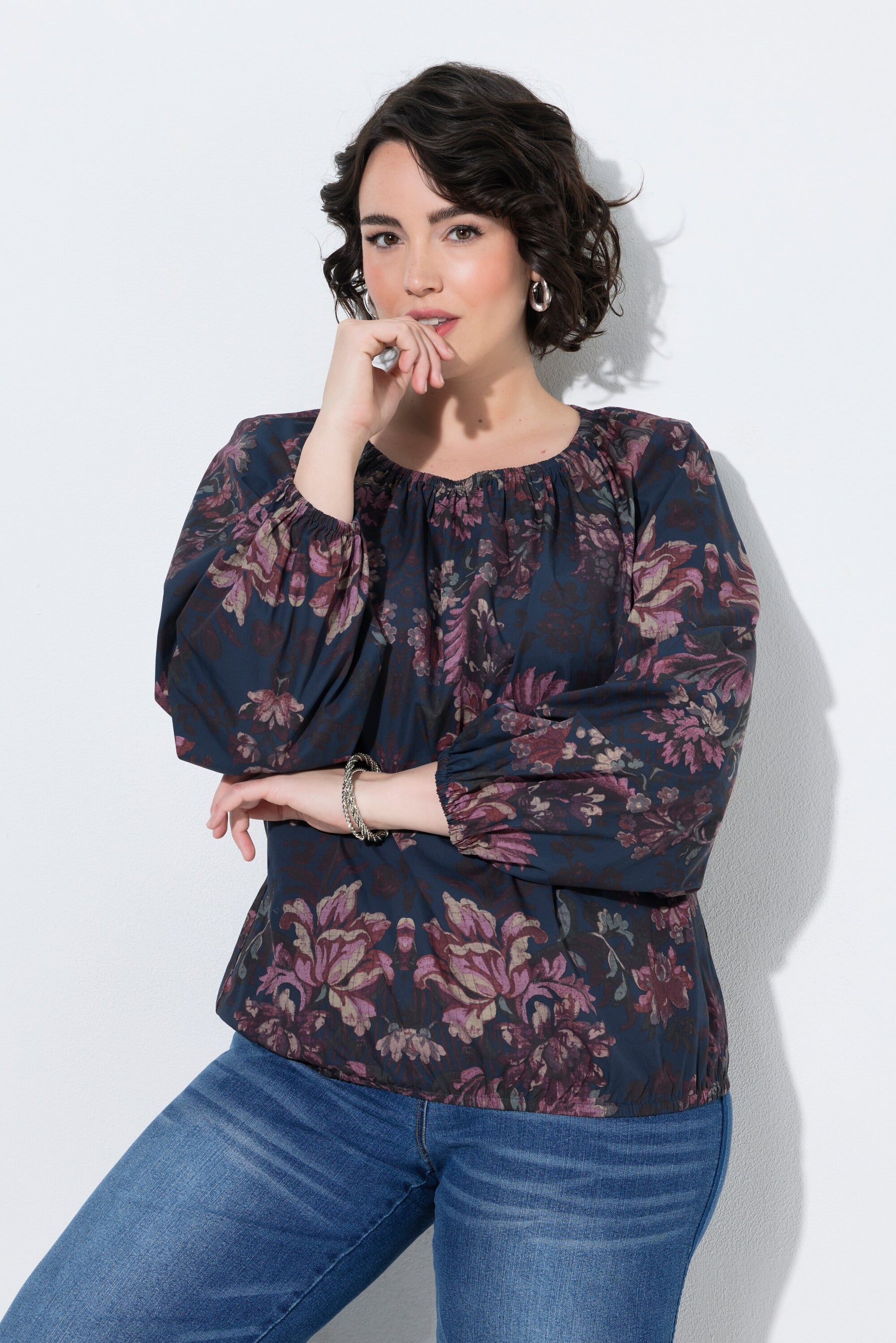 A woman wearing the Ulla Popken Floral Damask Blouse in Navy, a dark navy floral blouse with purple and pink tones, paired with blue jeans, posing against a light gray studio background.