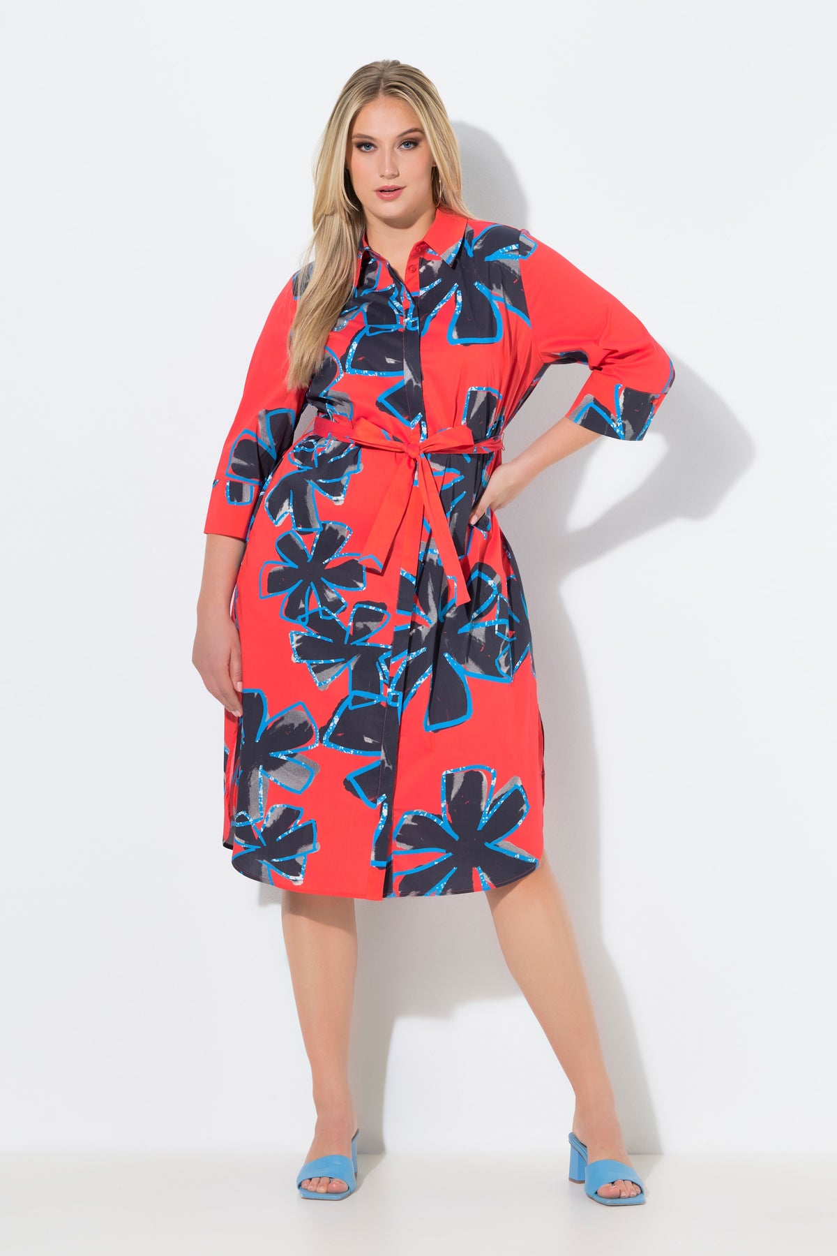 Woman wearing the Ulla Popken Floral Shirt Dress in Red, a red dress with blue floral pattern. This dress has a shirt collar, 3/4 length sleeves and a midi length, with a removable tie belt at the waist on a white background