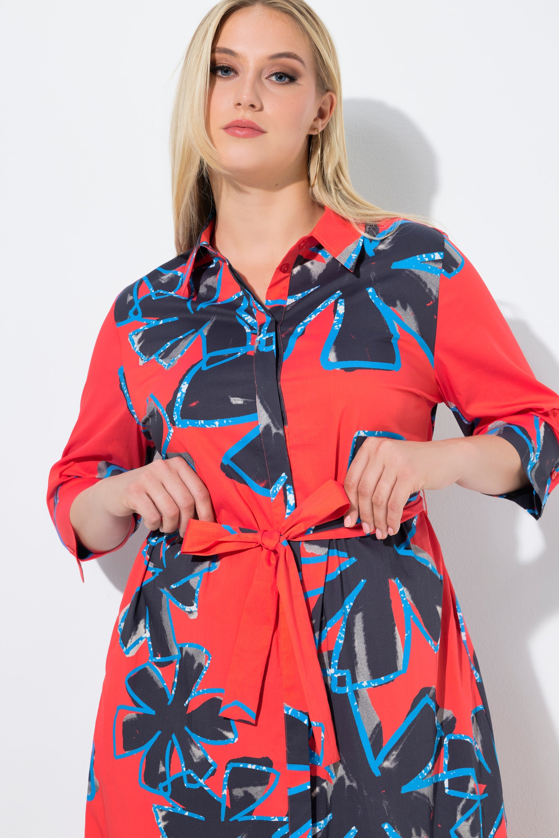 Woman wearing the Ulla Popken Floral Shirt Dress in Red, a red dress with blue floral pattern. This dress has a shirt collar, 3/4 length sleeves and a midi length, with a removable tie belt at the waist on a white background   