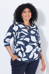 A woman with curly black hair wearing the Ulla Popken Keyhole Leaf Print Blouse in Navy, a navy blue, white, and light blue abstract patterned blouse and dark pants, standing against a white studio background.