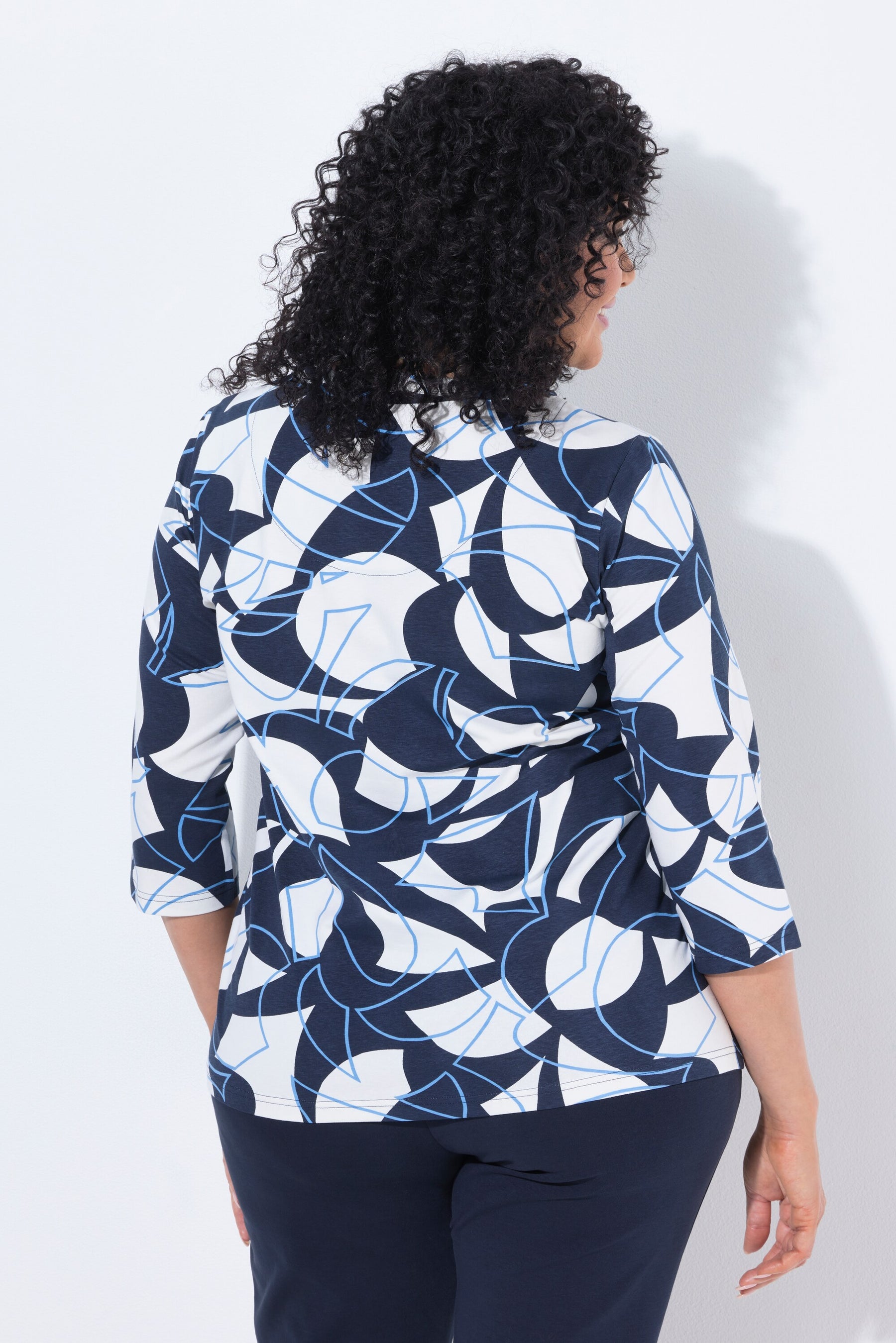 A woman with curly black hair is shown from the back wearing the Ulla Popken Keyhole Leaf Print Blouse in Navy, a navy blue, white, and light blue abstract patterned blouse with three-quarter sleeves, paired with navy pants, against a plain white background.
