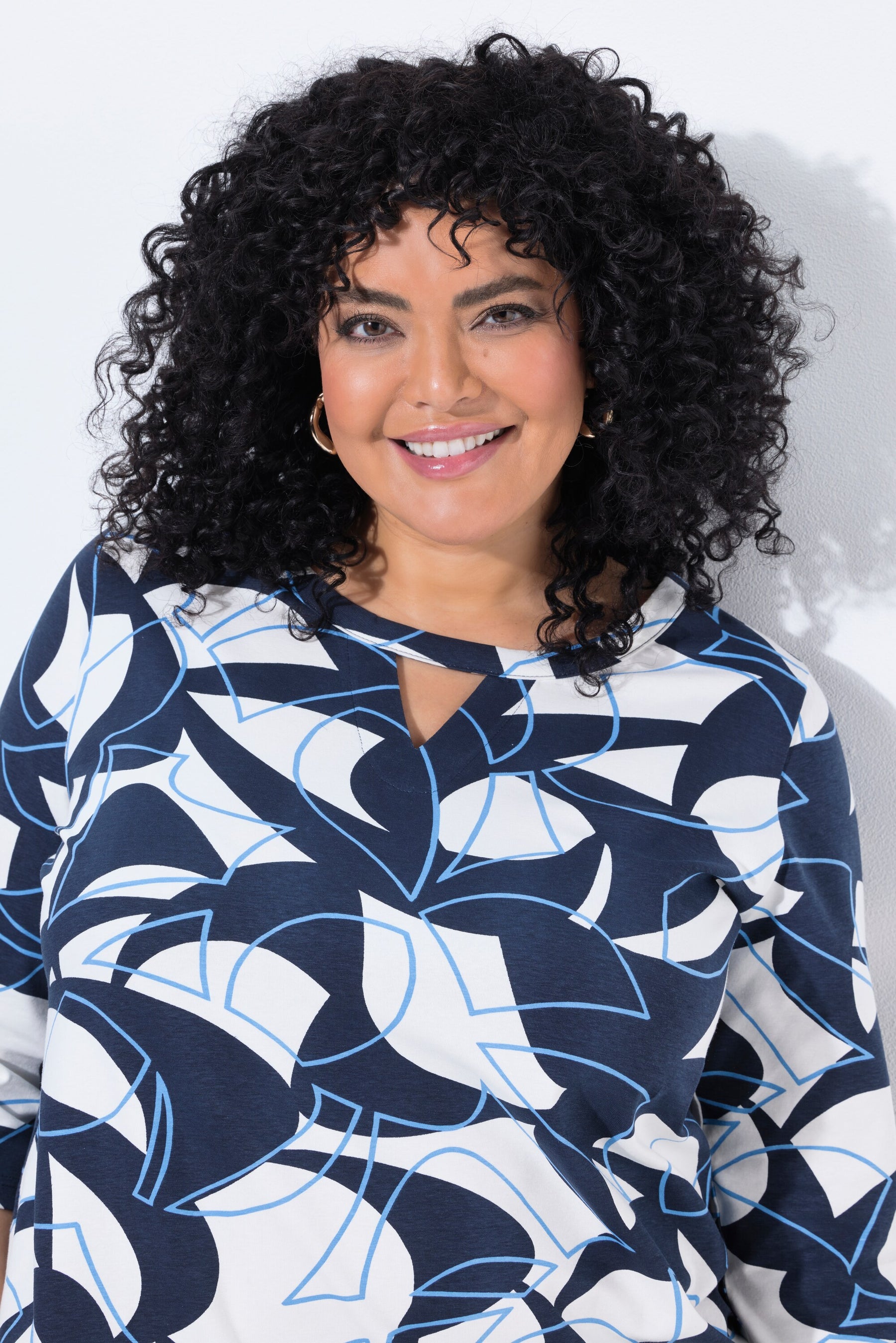 A smiling woman with voluminous curly black hair wearing the Ulla Popken Keyhole Leaf Print Blouse in Navy, a navy, white, and light blue abstract patterned long-sleeve top, photographed against a plain white background.