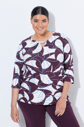 A woman modeling the Ulla Popken Keyhole Leaf Print Blouse in Wine, a three-quarter sleeve top with a wine, white, and light blue abstract pattern, paired with matching purple pants, against a light gray studio background.