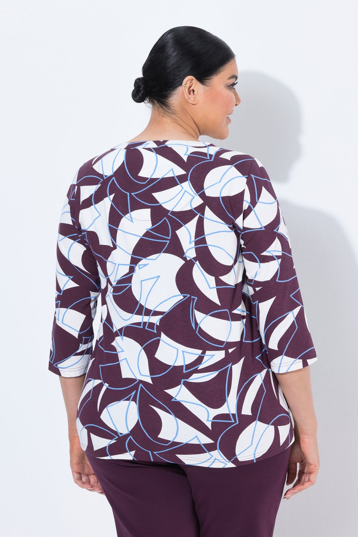 A woman shown from the back wearing the Ulla Popken Keyhole Leaf Print Blouse in Wine, a three-quarter sleeve top with an abstract geometric pattern in wine, white, and light blue, paired with solid dark purple pants, against a plain white background.