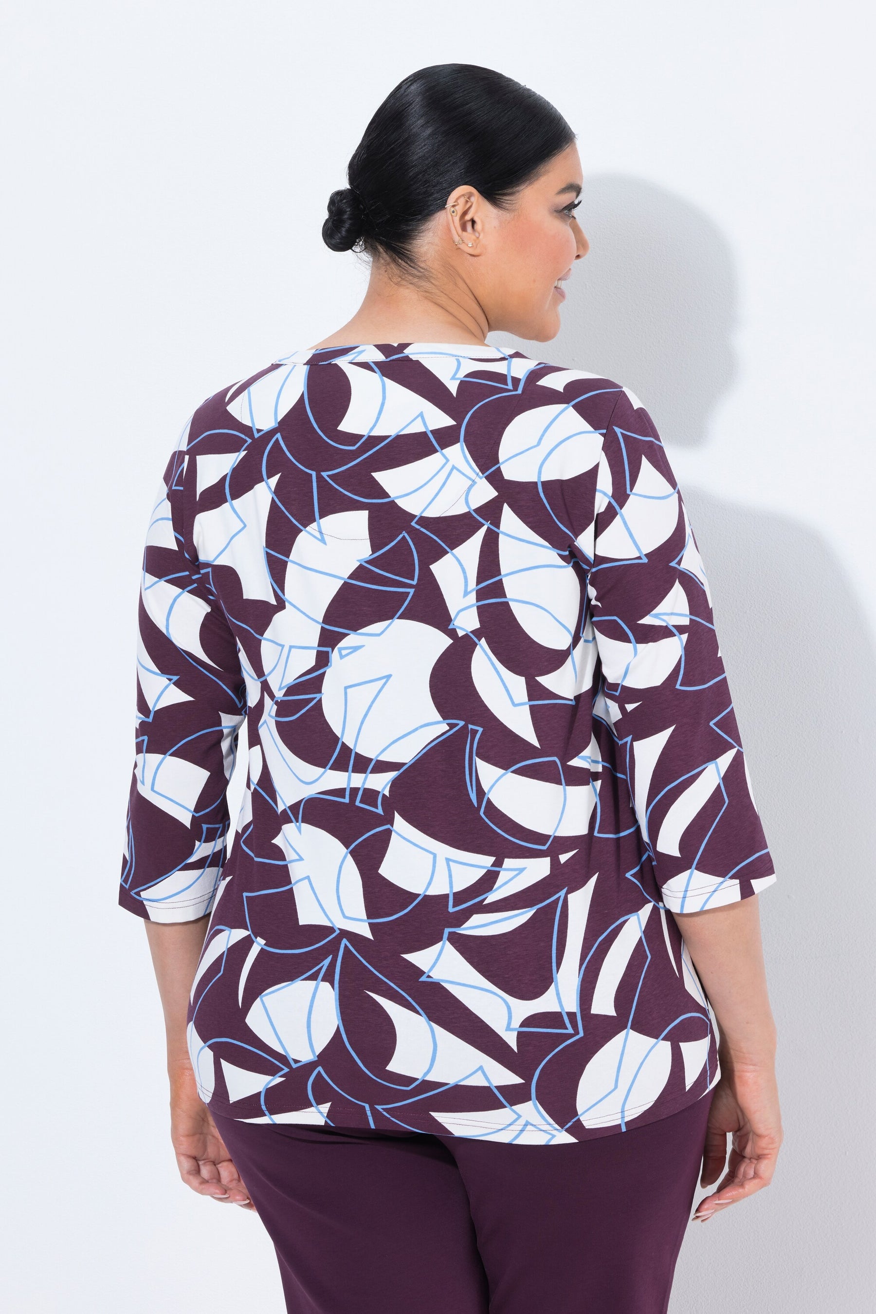 A woman shown from the back wearing the Ulla Popken Keyhole Leaf Print Blouse in Wine, a three-quarter sleeve top with an abstract geometric pattern in wine, white, and light blue, paired with solid dark purple pants, against a plain white background.