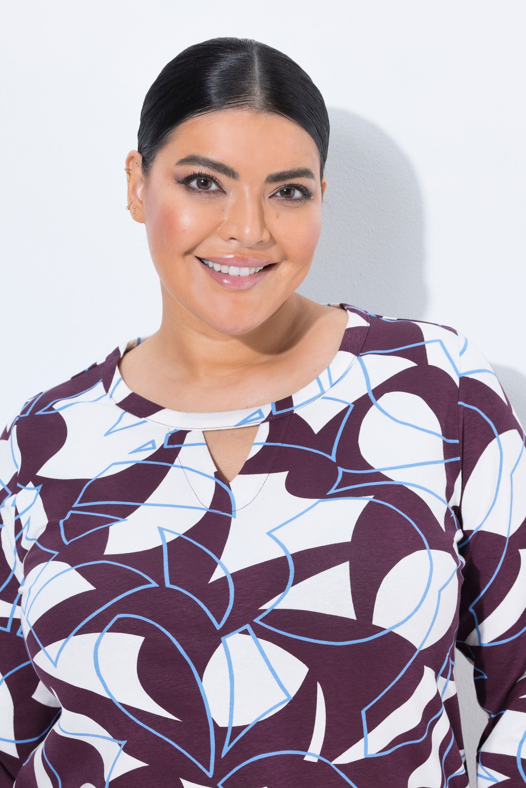 A woman with dark hair pulled back, wearing the Ulla Popken Keyhole Leaf Print Blouse in Wine, a long-sleeve top with a white, wine, and light blue abstract pattern, photographed against a plain white background.