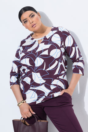 A woman models the Ulla Popken Keyhole Leaf Print Blouse in Wine, a 3/4 sleeve blouse with a bold abstract pattern in white, wine, and light blue, paired with matching purple trousers and holding a wine handbag against a light gray wall.