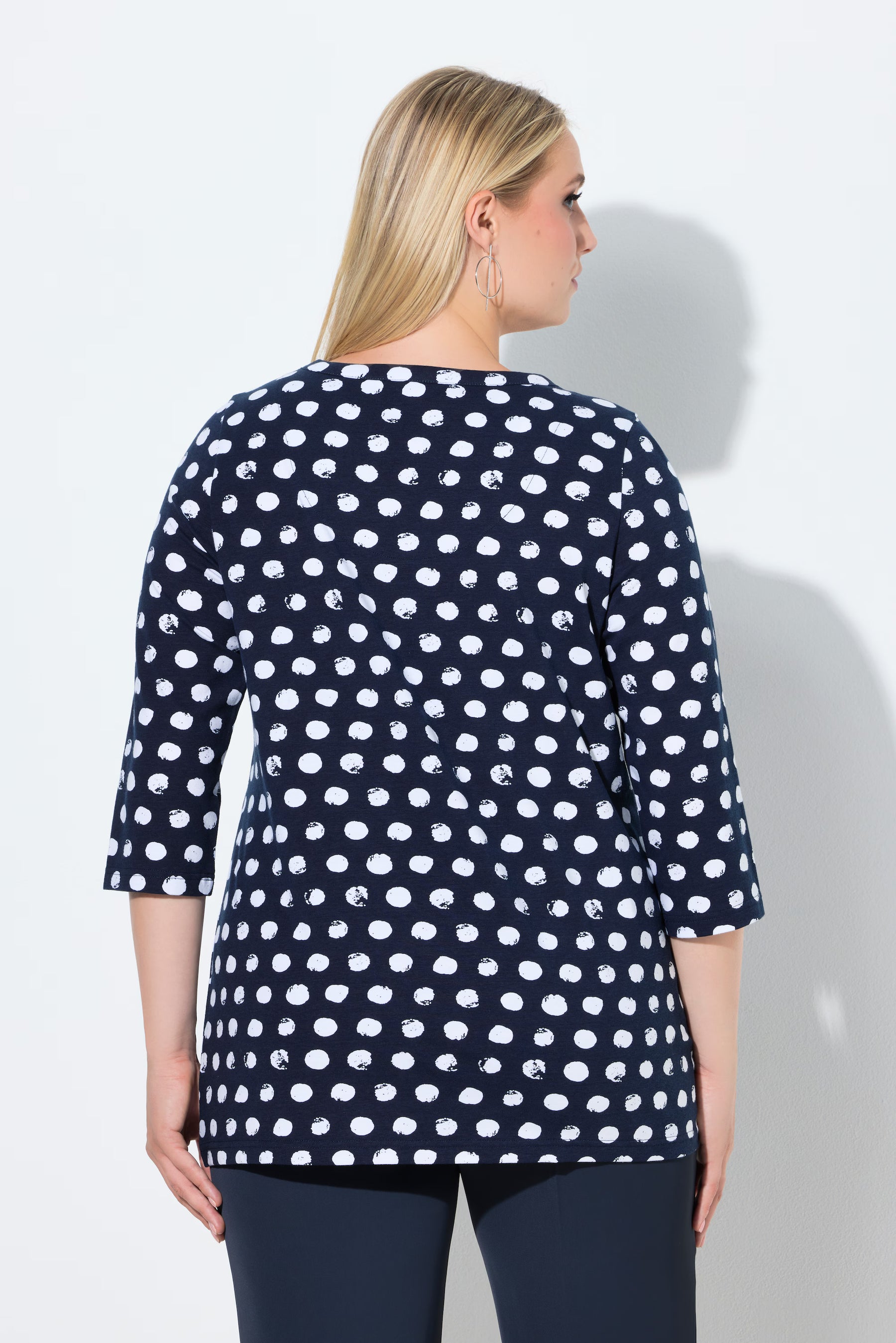 Woman wearing the Ulla Popken Polka Dot Keyhole Top in Navy, a navy blue top with white polka dots, 3/4 length sleeves and a round neckline with a keyhole cut out against a light gray background