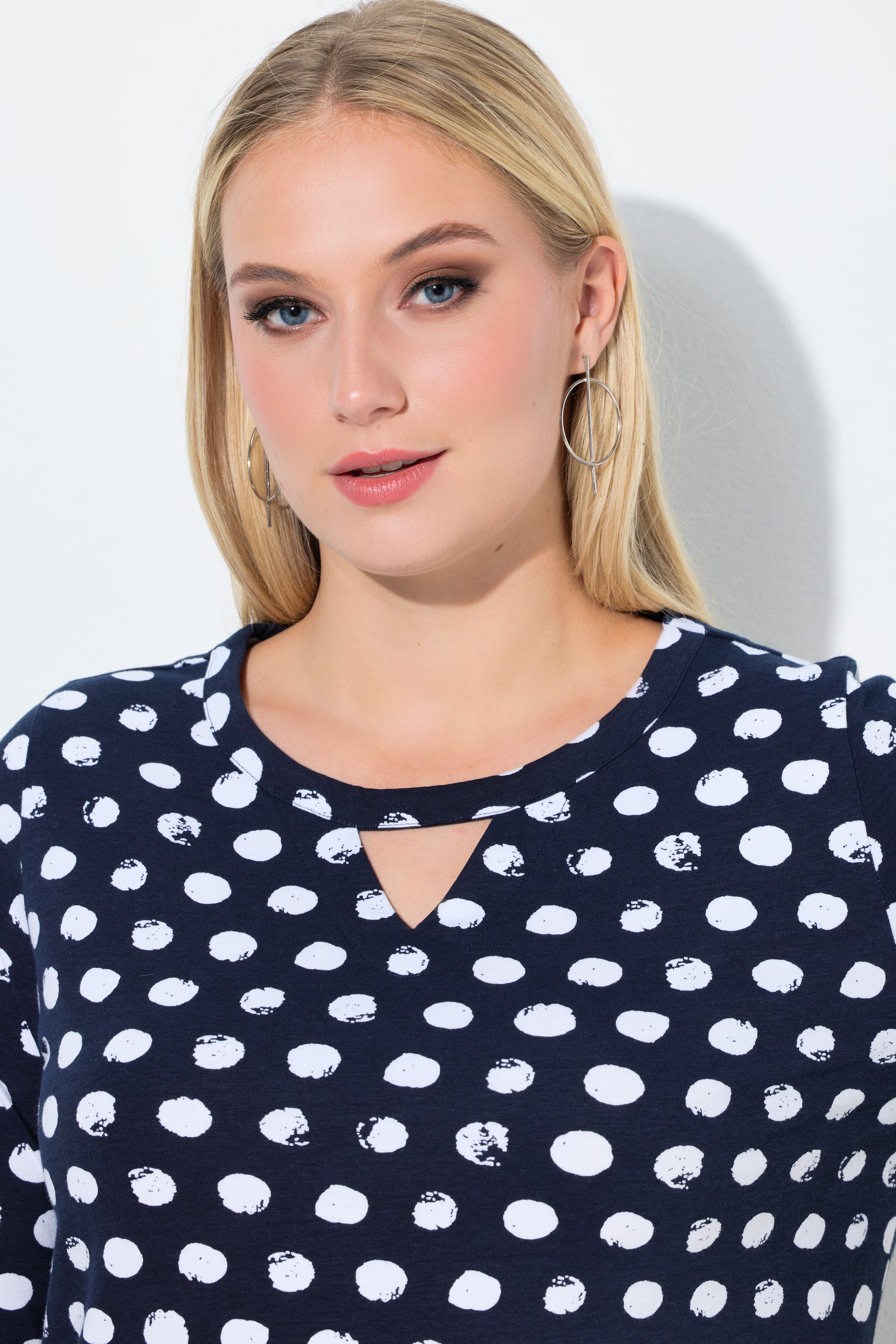 Woman wearing the Ulla Popken Polka Dot Keyhole Top in Navy, a top with a round neckline with a keyhole cut out and in navy with white polka dot pattern against a plain background