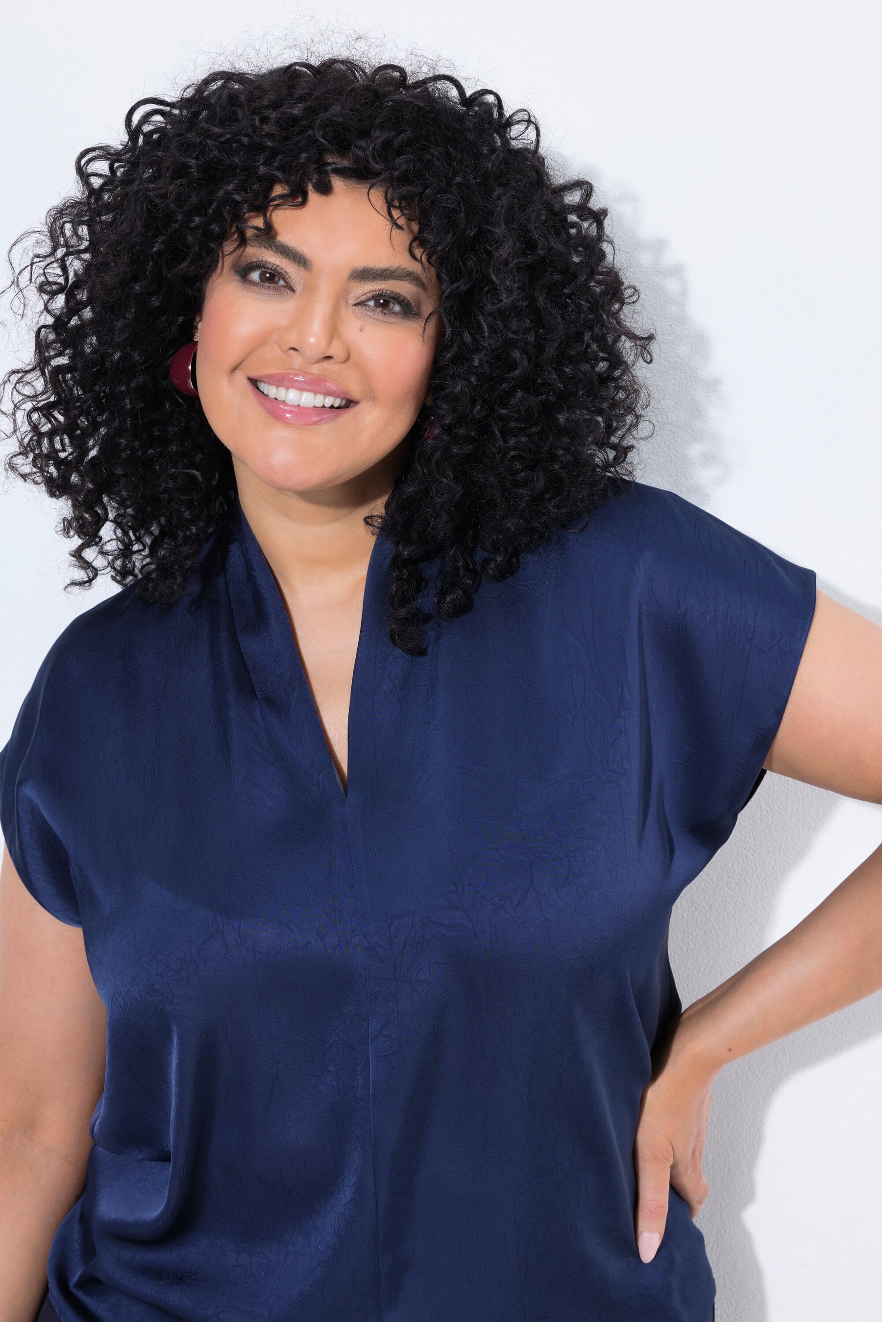 A woman with voluminous curly black hair wearing the Ulla Popken Satin Blouse in Navy, a short-sleeved navy blue satin blouse, posing against a light gray studio background.