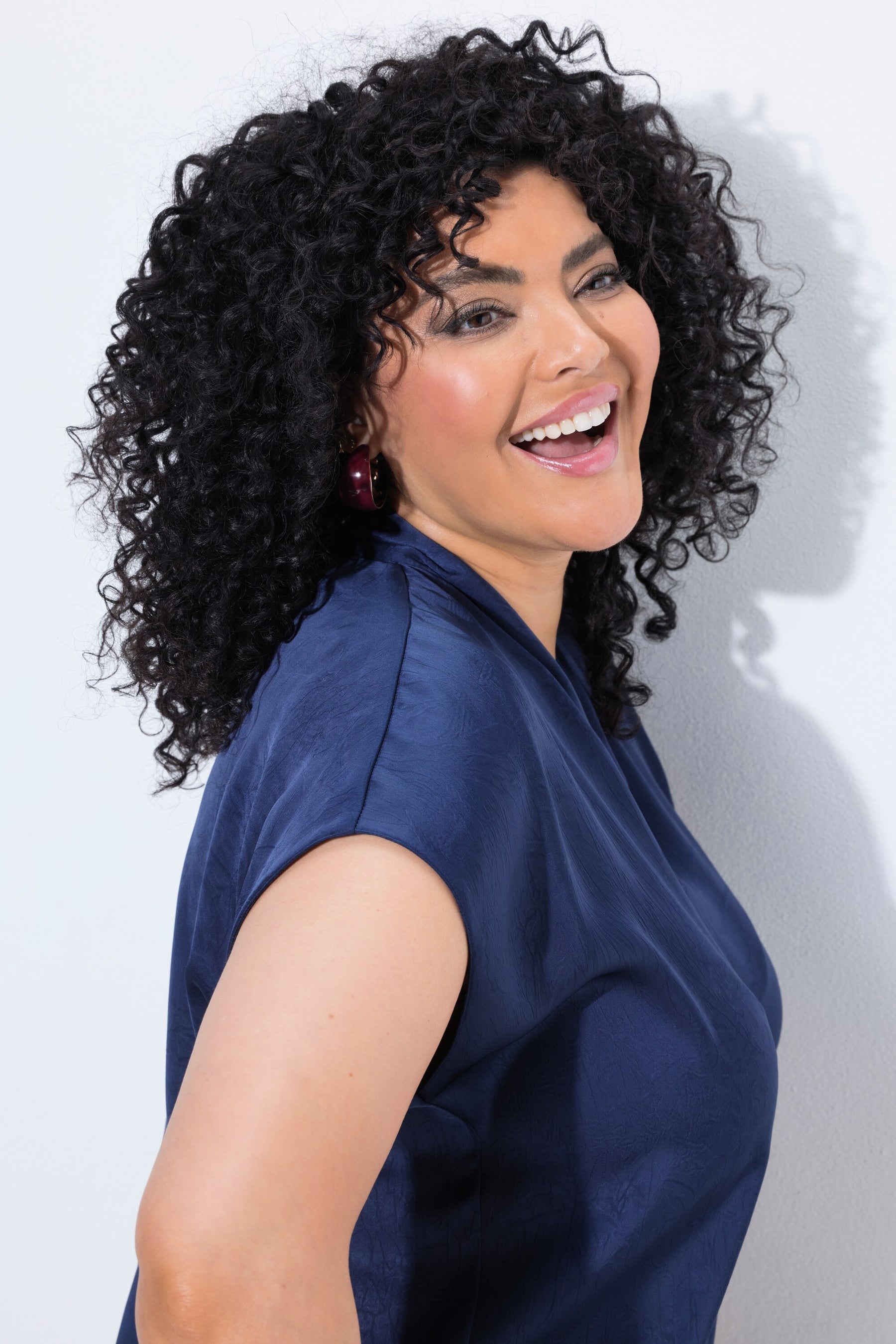 A smiling woman with voluminous curly black hair wearing the Ulla Popken Satin Blouse in Navy, a navy blue satin top, shown in a side profile against a light gray wall.