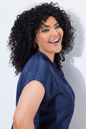 A smiling woman with voluminous curly black hair wearing the Ulla Popken Satin Blouse in Navy, a navy blue satin top, shown in a side profile against a light gray wall.