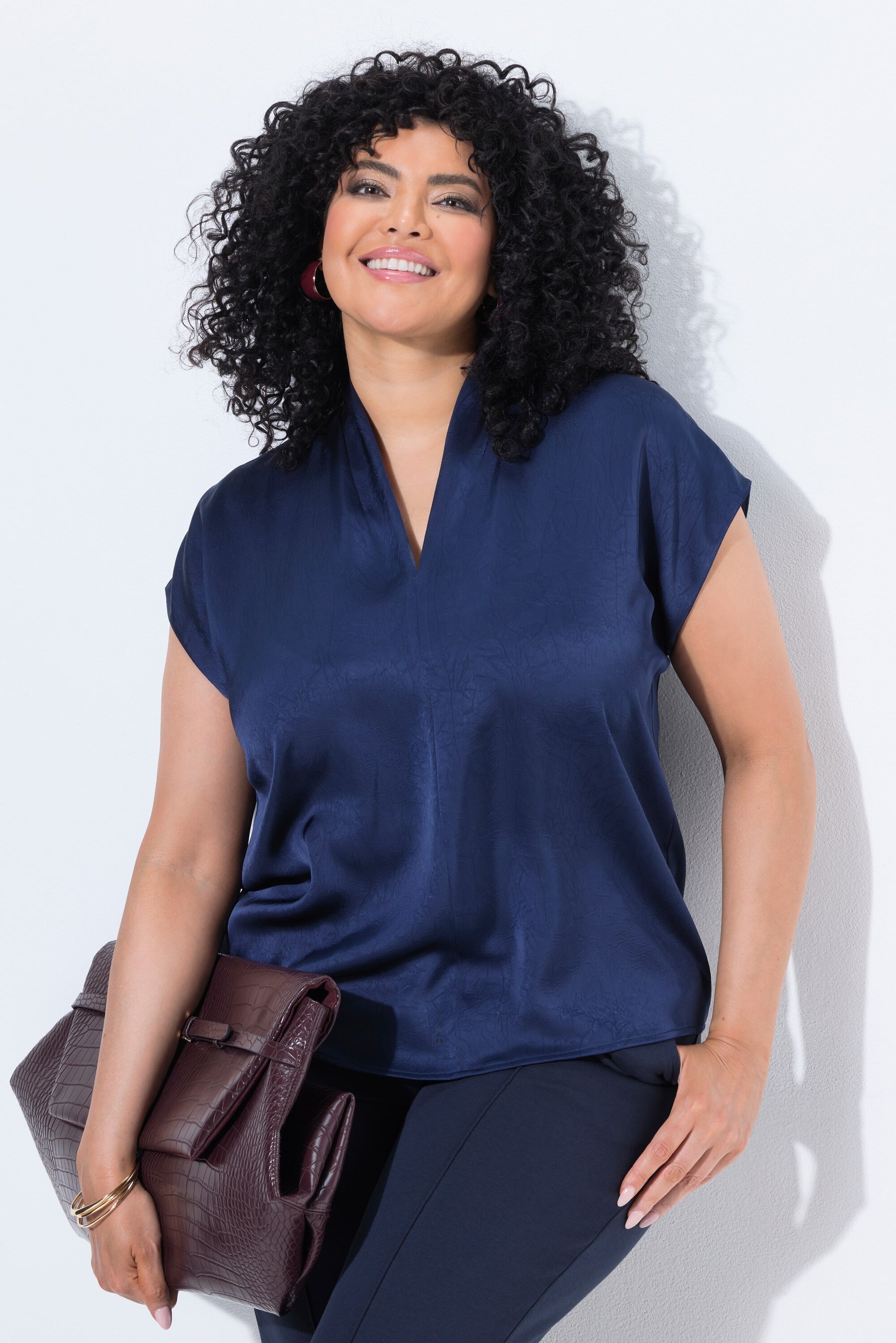 A woman with curly black hair wearing the Ulla Popken Satin Blouse in Navy, a navy blue short-sleeve satin top and dark pants, holding a brown textured clutch bag against a plain white background.