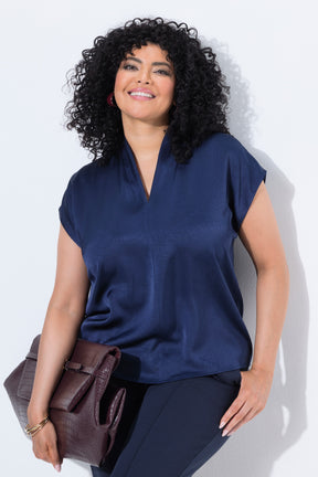 A woman with curly black hair wearing the Ulla Popken Satin Blouse in Navy, a navy blue short-sleeve satin top and dark pants, holding a brown textured clutch bag against a plain white background.