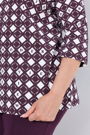 A close-up of a person wearing the Ulla Popken Wine Print Blouse, a short-sleeve top with a repeating geometric diamond pattern in dark plum and white, paired with matching plum bottoms. The person's hand is visible at the side hem.