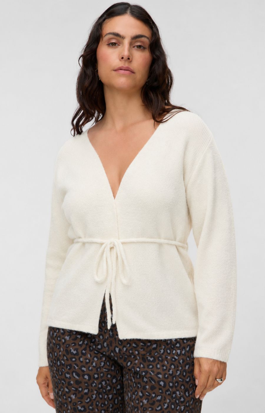 A woman wearing the Vero Moda Curve Cora Cardigan in Cream, a cream-colored knit wrap cardigan with a tie waist, paired with dark patterned pants, against a plain light gray background. This image was created using AI.