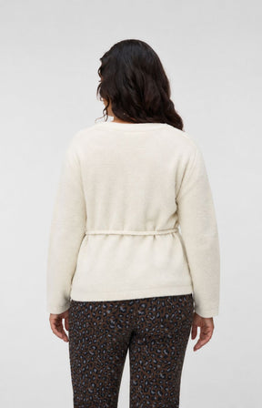 A woman viewed from the back wearing the Vero Moda Curve Cora Cardigan in Cream, a cream-colored knit cardigan with a tie waist detail and dark patterned pants, set against a plain light gray studio background. This image was created using AI.