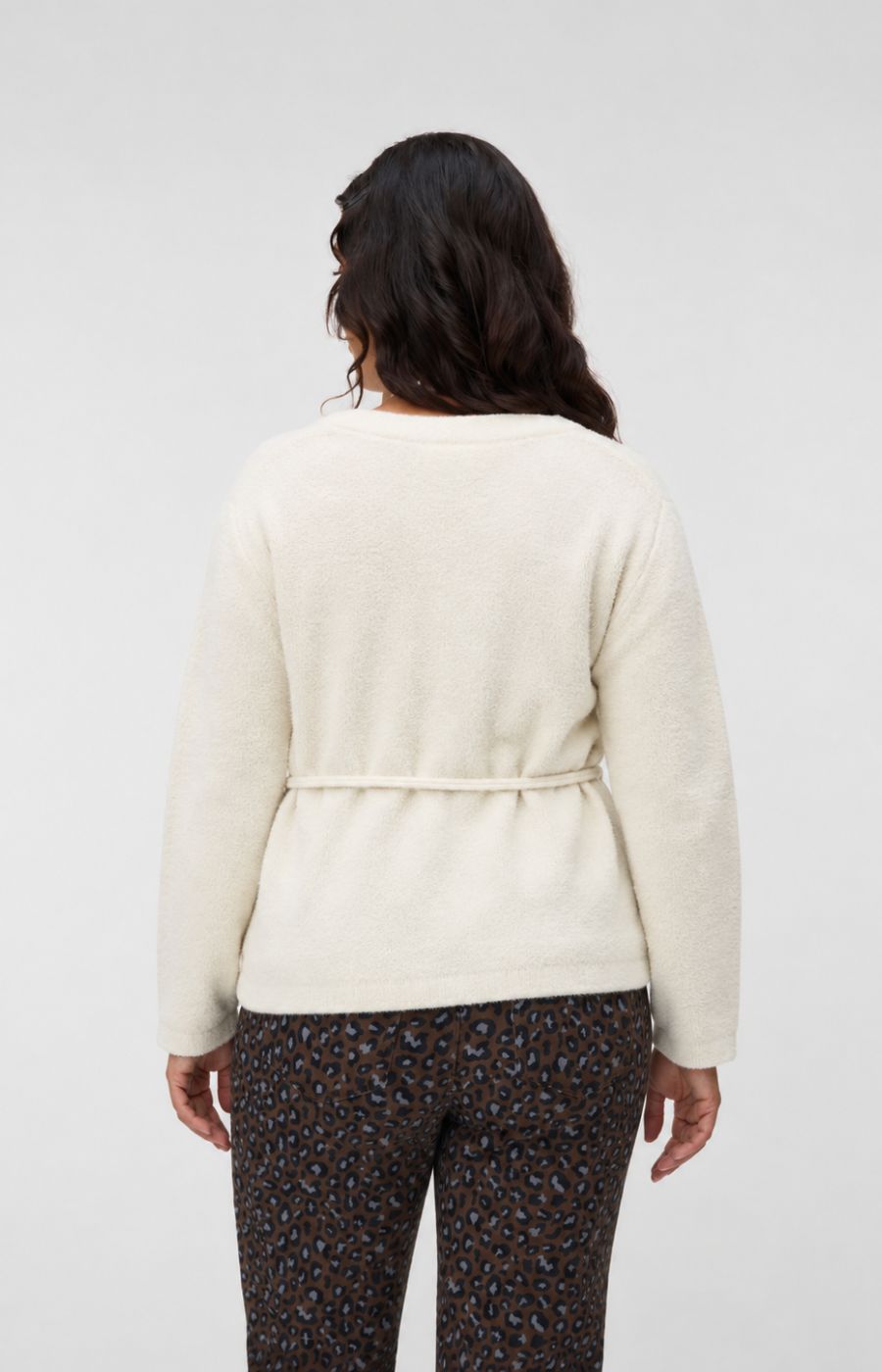 A woman viewed from the back wearing the Vero Moda Curve Cora Cardigan in Cream, a cream-colored knit cardigan with a tie waist detail and dark patterned pants, set against a plain light gray studio background. This image was created using AI.