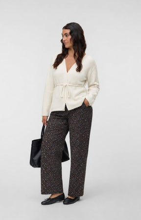 A woman modeling the Vero Moda Curve Cora Cardigan in Cream, a cream wrap cardigan with a tie waist, paired with dark patterned wide-leg trousers, black flats, and holding a black tote bag, against a light gray studio background. This image was created using AI.