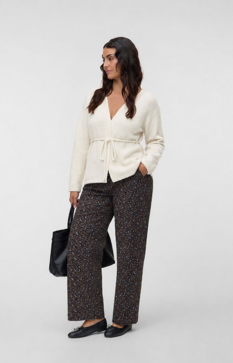 A woman modeling the Vero Moda Curve Cora Cardigan in Cream, a cream wrap cardigan with a tie waist, paired with dark patterned wide-leg trousers, black flats, and holding a black tote bag, against a light gray studio background. This image was created using AI.