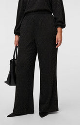A model wearing the Vero Moda Curve Kali Trousers in Black, a straight leg trouser with a black long-sleeve top with a subtle glitter texture, holding a black leather handbag against a light gray studio background. This image was created using AI.