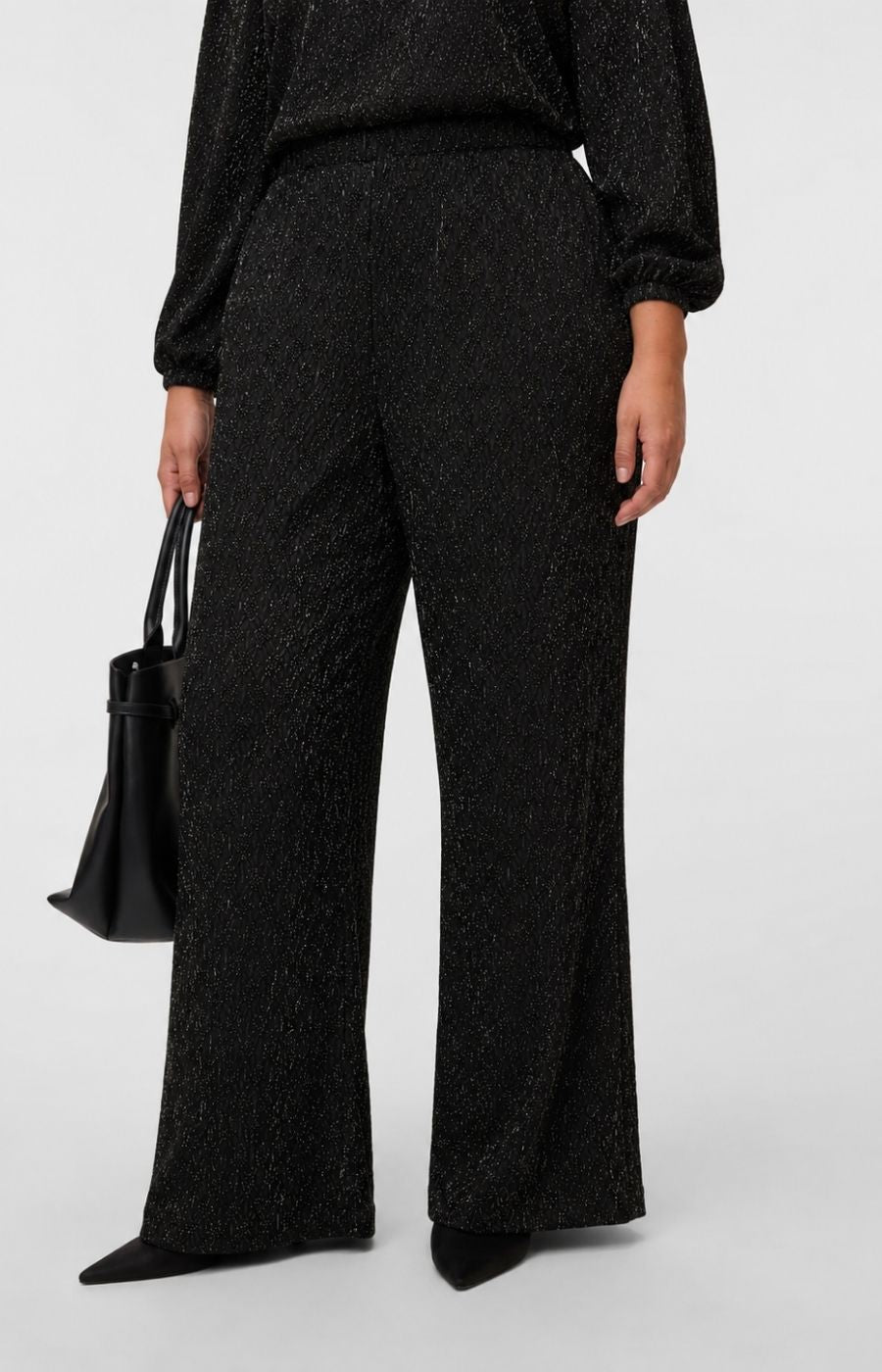 A model wearing the Vero Moda Curve Kali Trousers in Black, a straight leg trouser with a black long-sleeve top with a subtle glitter texture, holding a black leather handbag against a light gray studio background. This image was created using AI.