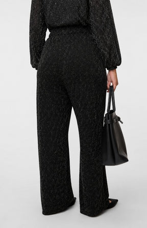 A model shown from the back wearing the Vero Moda Curve Kali Trousers in Black,a black textured, sparkly trousers with a straight leg, worn with the matching long sleeve top and holding a black leather handbag against a light gray studio background. This image was created using AI.