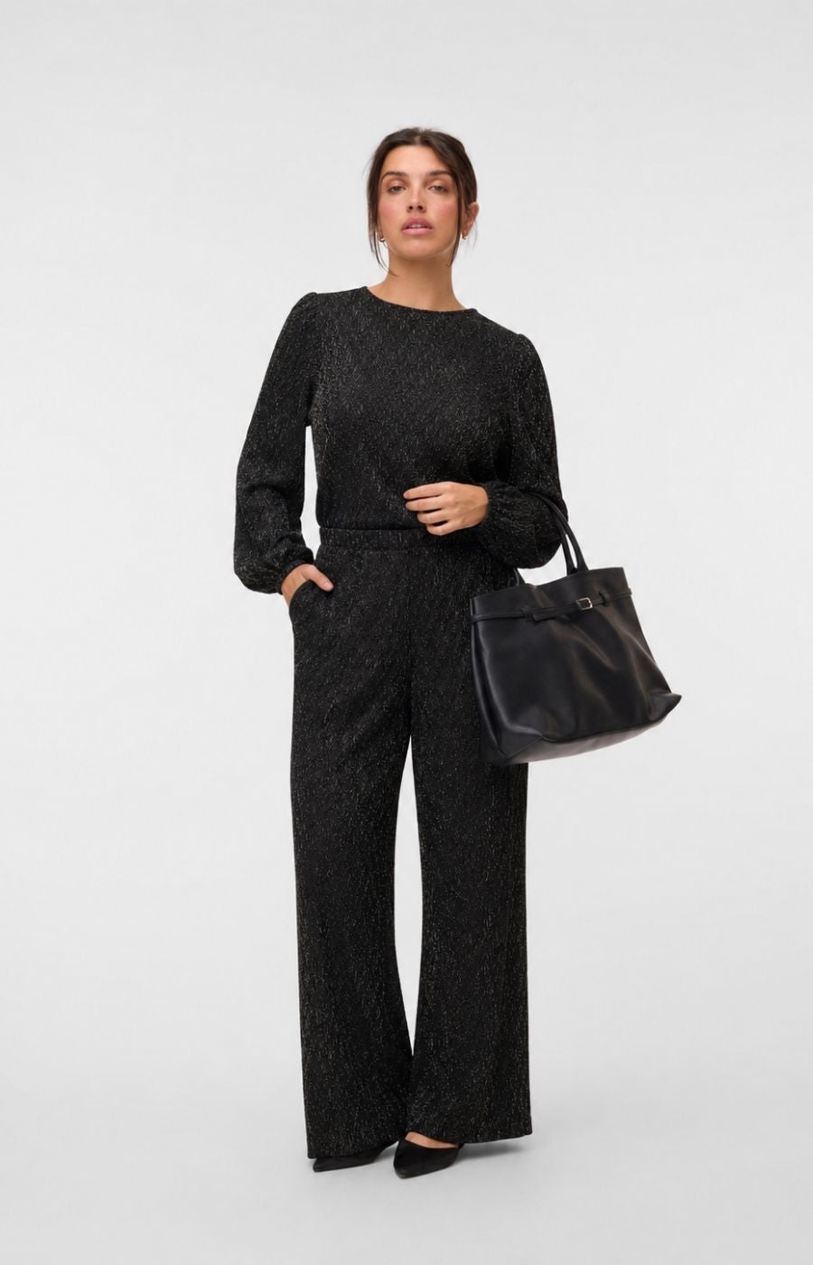A woman modeling the Vero Moda Curve Kali Trousers in Black, a textured, sparkling, black straight leg trousers, worn with the matching top,  holding a black leather tote bag against a light gray studio background. This image was created using AI. 