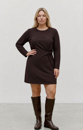 A woman modeling the Vero Moda Curve Kanva Dress in Chocolate, a dark brown long-sleeve knit dress with a twisted waist detail, paired with matching dark brown knee-high boots, against a neutral studio background. This image was created using AI.