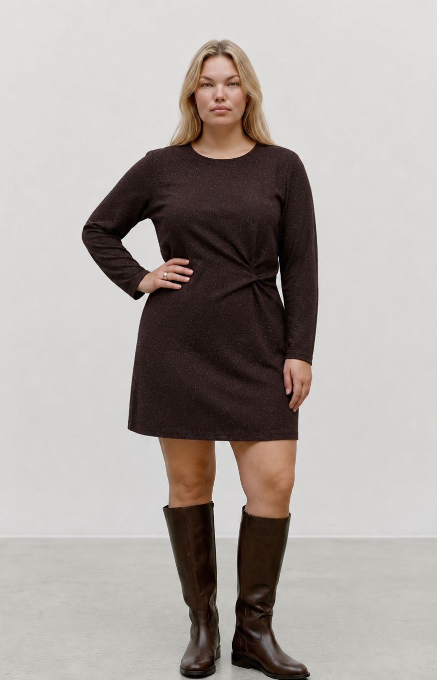 A woman modeling the Vero Moda Curve Kanva Dress in Chocolate, a dark brown long-sleeve knit dress with a twisted waist detail, paired with matching dark brown knee-high boots, against a neutral studio background. This image was created using AI.