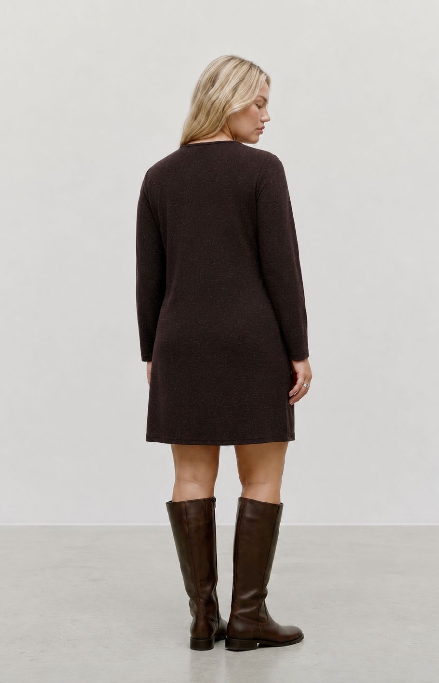 A woman stands with her back turned wearing the Vero Moda Curve Kanva Dress in Chocolate, a dark brown long-sleeve knit dress and matching dark brown knee-high boots against a plain light gray studio background. This image was created using AI.