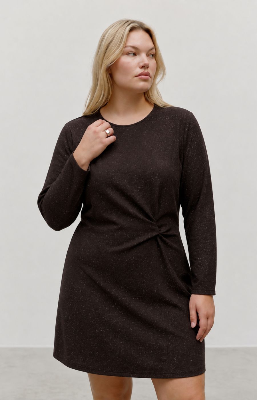 A woman modeling the Vero Moda Curve Kanva Dress in Chocolate, a dark brown long-sleeve knit dress with a gathered twist detail at the waist, set against a plain light gray studio background. This image was created using AI.