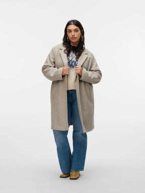 Woman wearing a beige coat over a patterned sweater, blue jeans, and brown shoes on a white background