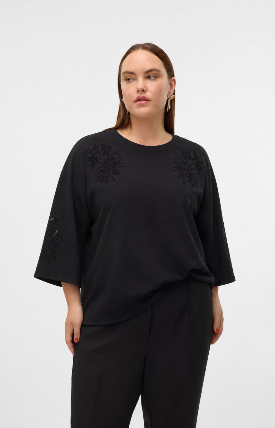 A woman modeling the Vero Moda Curve Nia Top in Black, a black short-sleeve top with tonal floral embroidery on the shoulders and sleeves, paired with black trousers, against a plain light gray studio background. This image was created using AI.