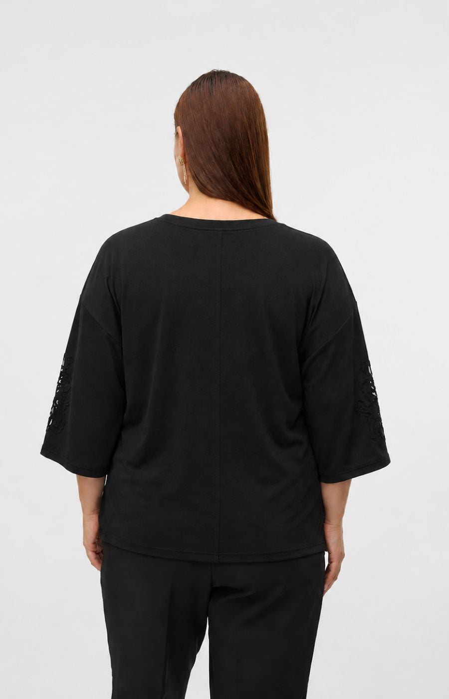 A woman viewed from the back wearing the Vero Moda Curve Nia Top in Black, a black 1/2 sleeve top with lace-up detailing on the sleeves and black pants, set against a plain light gray studio background. This image was created using AI.