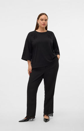 A woman modeling the Vero Moda Curve Nia Top in Black, a black 1/2 sleeve top with sequin detailing on the shoulders and  wide-leg trousers, photographed against a plain light gray studio background. This image was created using AI.
