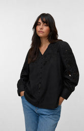 A woman wearing the Vero Moda Curve Olive Shirt in Black, a black long-sleeve blouse with lace detailing on the chest and sleeves, paired with blue jeans, photographed against a light gray studio background. This image was created using AI.