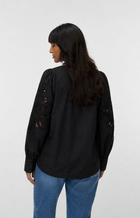 A woman shown from the back wearing the Vero Moda Curve Olive Shirt in Black, a black long-sleeve blouse with distressed cutout details on the sleeves, paired with blue jeans, against a plain light gray studio background. This image was created using AI