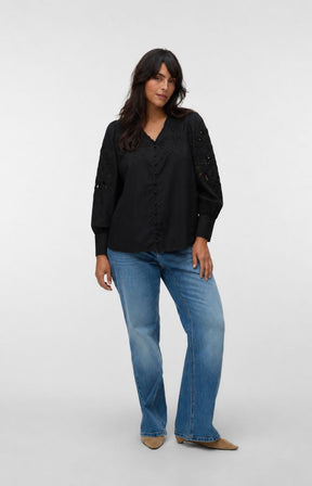 A woman modeling the Vero Moda Curve Olive Shirt in Black, a black long-sleeve blouse with lace detailing on the sleeves and front, paired with blue jeans and tan pointed-toe flats, against a light gray studio background. This image was created using AI.