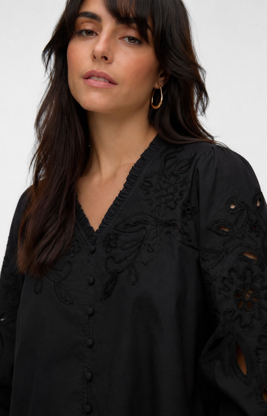 A close-up of a woman wearing the Vero Moda Curve Olive Shirt in Black, a black embroidered blouse with cutout details on the sleeves, set against a plain light gray background. This image was created using AI.