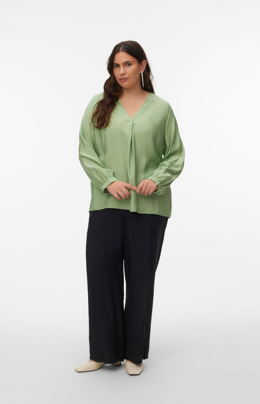 Woman wearing a green blouse with a V neckline, inverted pleat and long sleeves with buttoned cuffs with a plain background