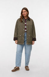 A woman modeling the Vero Moda Curve Wales Jacket in Grape Leaf, an olive green waxed cotton jacket with dark brown ribbed collar, cuffs, and pocket trims, worn over a striped shirt and blue jeans against a light gray studio background. This image was created using AI.
