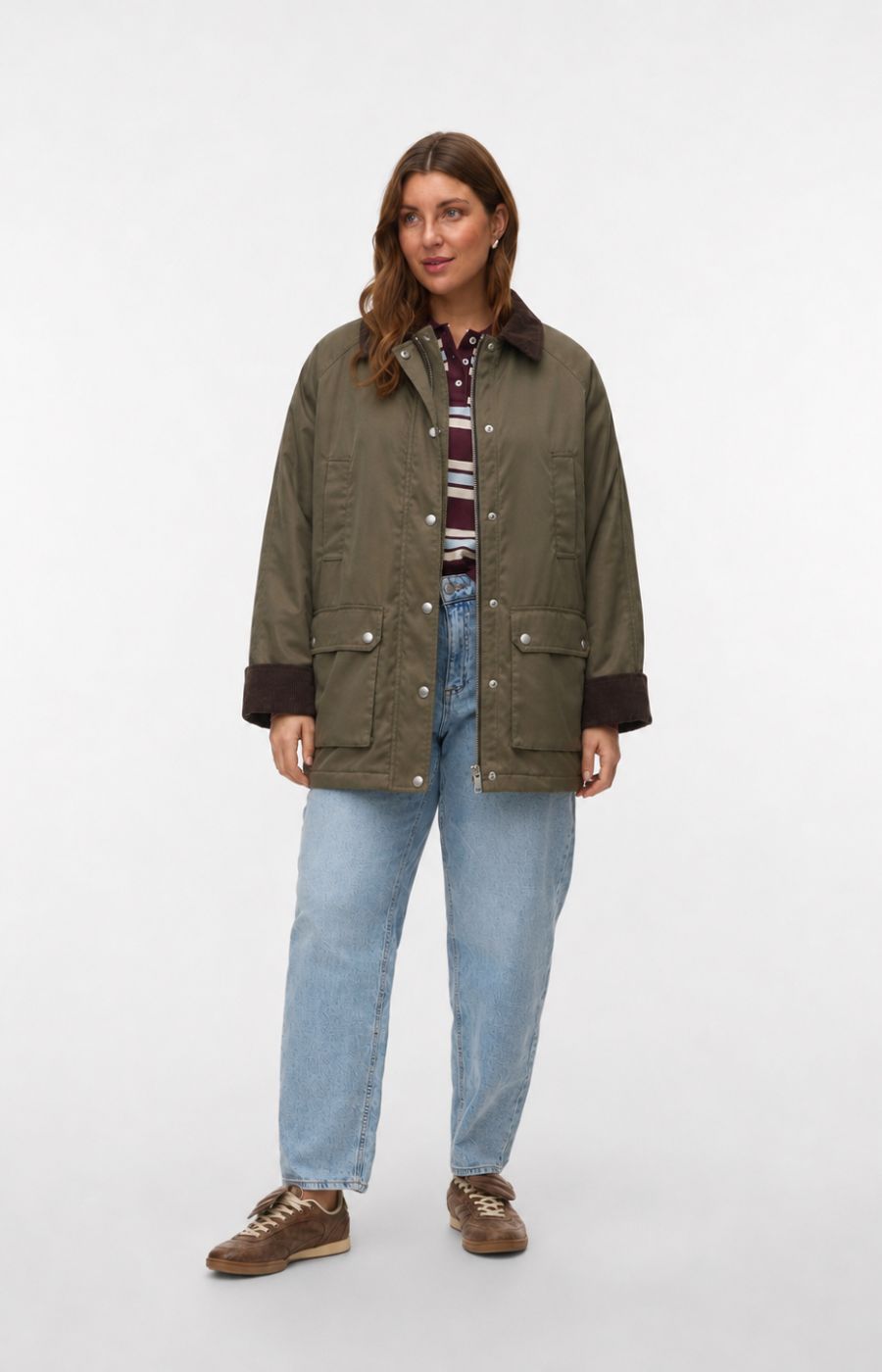 A woman modeling the Vero Moda Curve Wales Jacket in Grape Leaf, an olive green waxed cotton jacket with dark brown ribbed collar, cuffs, and pocket trims, worn over a striped shirt and blue jeans against a light gray studio background. This image was created using AI.