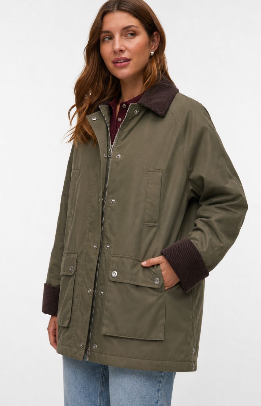 A woman wearing the Vero Moda Curve Wales Jacket in Grape Leaf,  an olive green jacket with dark brown collar, cuffs, and pocket trim, paired with blue jeans, against a plain white studio background. This image was created using AI.