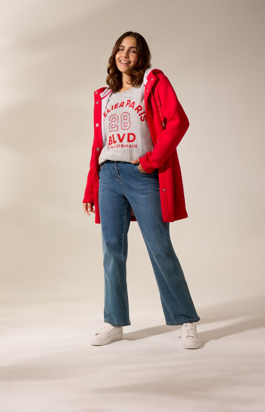 Woman wearing a red coat, white graphic t-shirt, blue jeans, and white sneakers on a beige background