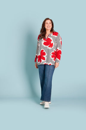 Lady wearing V neck blouse, long buttoned cuffs, white and navy stripe with a red floral print with denim jeans and white trainers