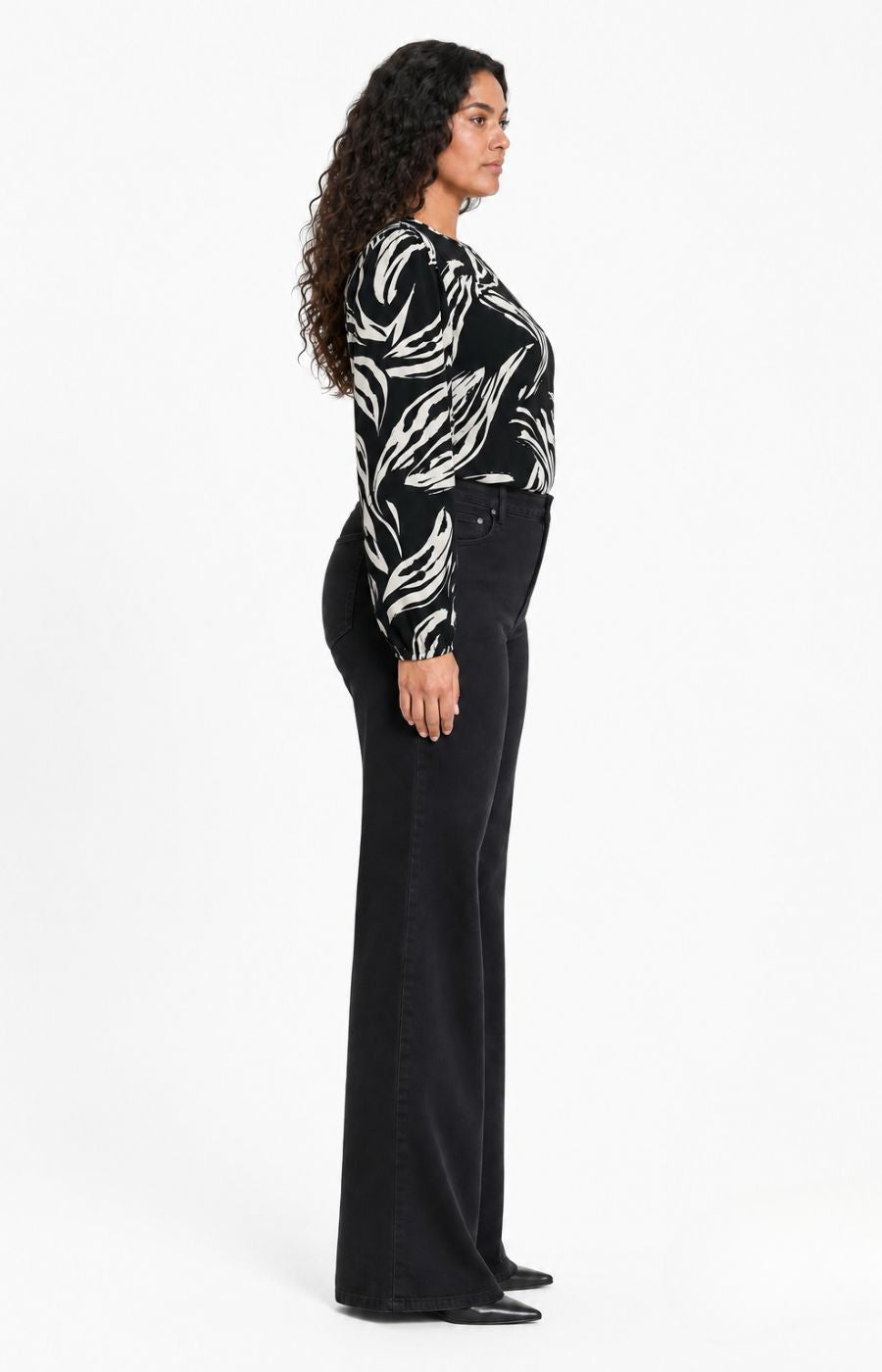 A woman shown in side profile wearing the Wasabi Juliette Top in Black, a black long-sleeve top with a white abstract leaf pattern and black wide-leg pants, standing against a plain white studio background. This image was created using AI