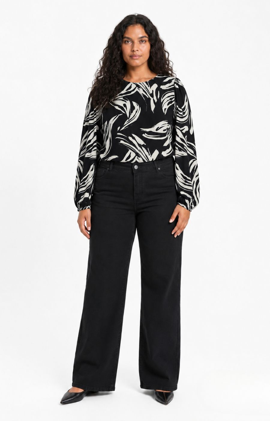 A woman modeling the Wasabi Juliette Top in Black, a black and white abstract patterned long-sleeve top paired with black wide-leg pants and black pointed shoes, photographed against a plain white studio background. This image was created using AI