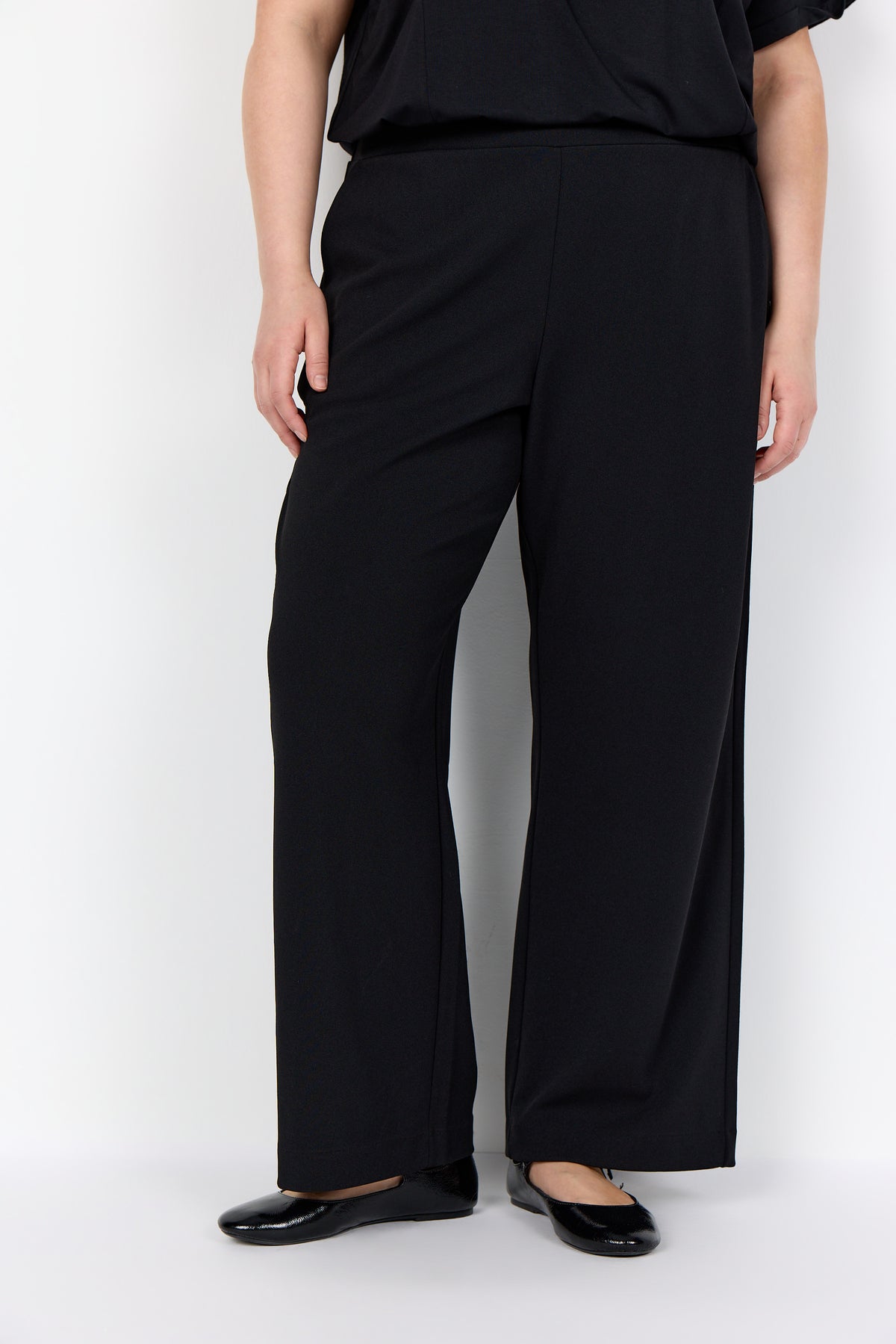 Person wearing Wasabi Kufa Trousers in Black, a full length straight leg, elasticated waistband and side pockets on a white background