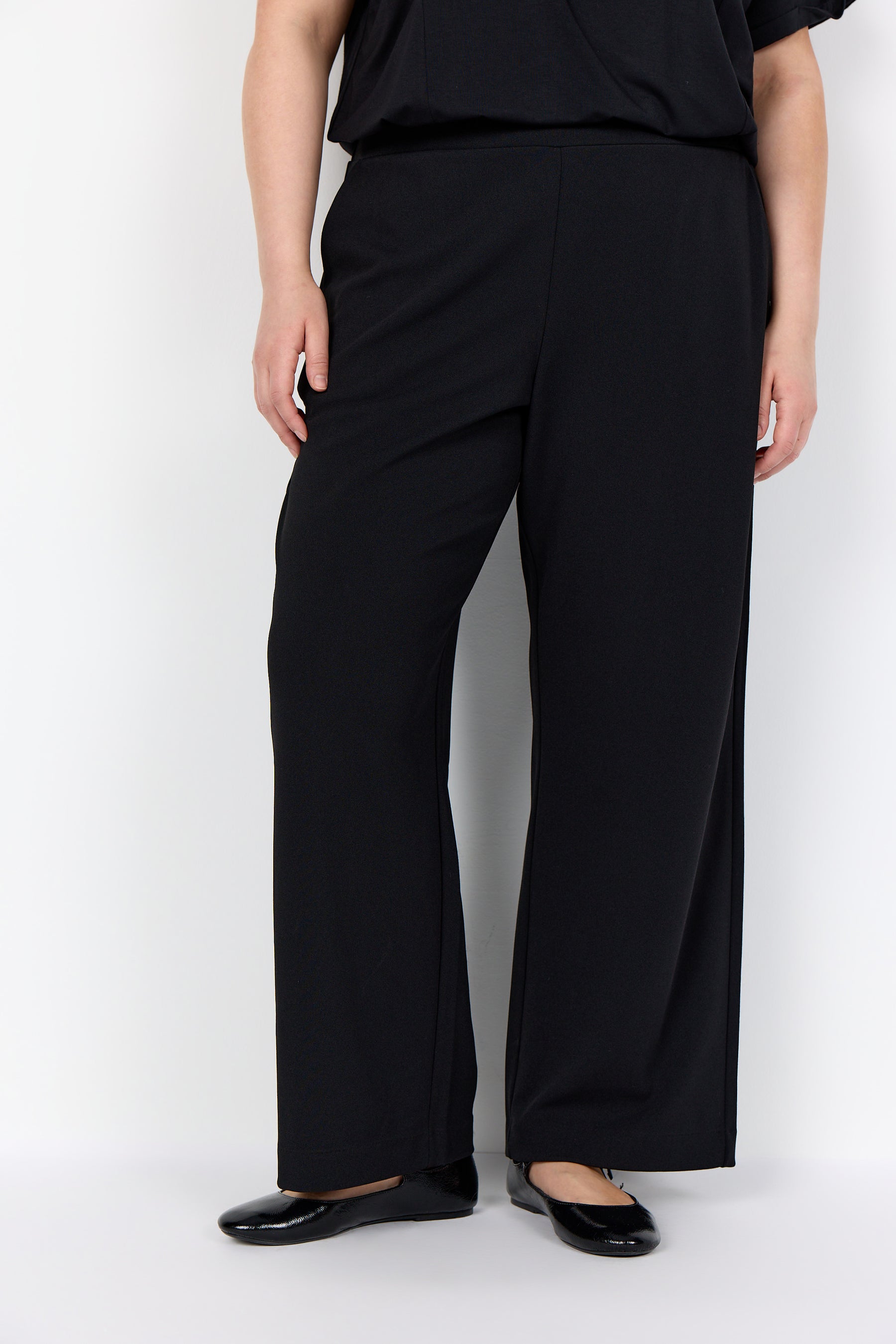 Person wearing Wasabi Kufa Trousers in Black, a full length straight leg, elasticated waistband and side pockets on a white background